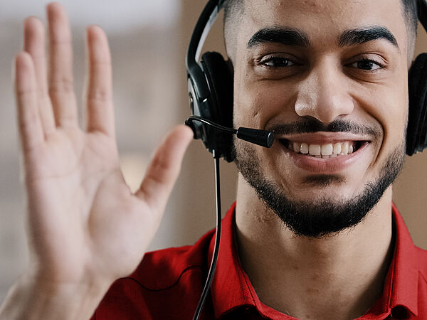 Ein Mann mit Headset lächelt in die Kamera und winkt dabei