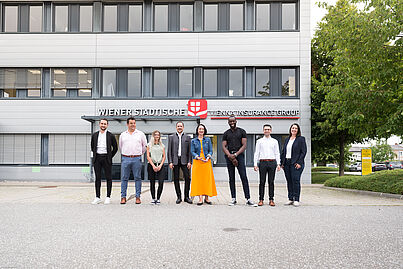 Mehrere Personen stehen vor einem Gebäude und blicken in die Kamera.