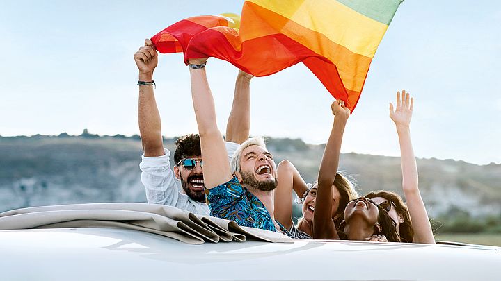 Eine Freundesgruppe fährt mit einem Van in den Urlaub und streckt voller Freude eine Fahne in den Himmel.