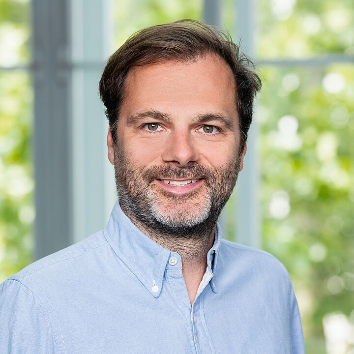 Ein Mann mit kurzen brauen Haaren und Dreitagebart trägt ein hellblaues Hemd. Er steht vor einem Fenster ins Grüne und lächelt.