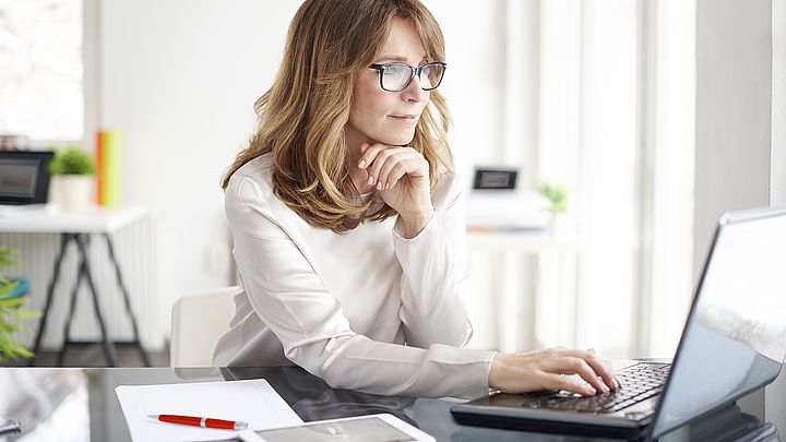 Eine Frau sitzt konzentriert im Büro am Schreibtisch vor ihrem Laptop.