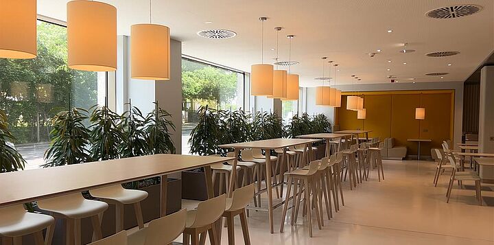 Kantine mit langen hohen Tischen, vielen Sesseln, Hängelampen und grünen Pflanzen.