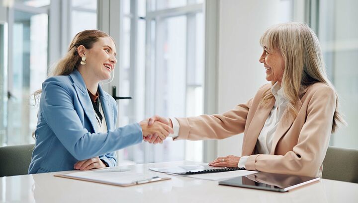 Zwei Frauen mit Sakko sitzen an einem Besprechungstisch gegenüber, schütteln sich die Hand und lächeln sich an.