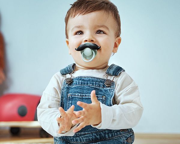 Baby mit Schnuller der aussieht wie ein Bart schaut in die Kamera.