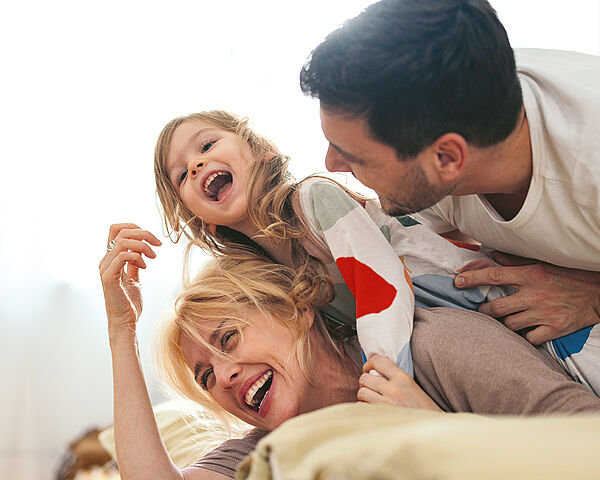 Ein lachendes Kind sitzt spielerisch auf den Schultern seiner Mutter, während der Vater sich von der Seite dazulehnt; alle drei wirken fröhlich und eng miteinander verbunden in einer hellen, entspannten Umgebung.