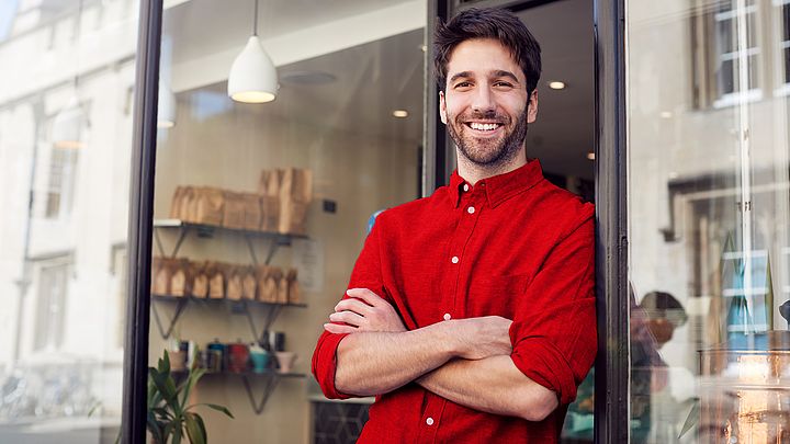 Ein junger Gründer eines Kaffeehauses steht vor seinem Geschäft und lächelt dabei voller Freude  