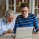 In einem Wohnzimmer sitzen zwei Männer nebeneinander am Tisch und schauen auf einen Laptop. Der ältere mit weißen Haaren und Bart trägt ein hellblaues Hemd, der junge eine Brille und einen dunkelblau-weiß-gestreiften Pullover.