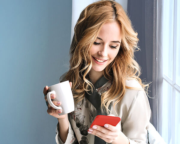 Eine junge Frau sitzt am Fensterbrett mit einer Tasse in der Hand und blickt fröhlich auf ihr Smartphone.