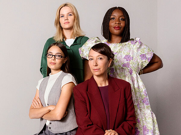 Vier Frauen posieren gemeinsam vor hellem Hintergrund. Im Vordergrund sitzt eine Frau in rotem Hosenanzug neben einer Frau mit Brille und grauer Weste. Dahinter stehen zwei weitere Frauen: links eine Blondine in grünem Oberteil, rechts eine Frau im geblümten Kleid.