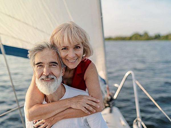 Ein glückliches Seniorenpaar steht auf einem Segelschiff am Meer. Die Frau umarmt den Mann von hinten.