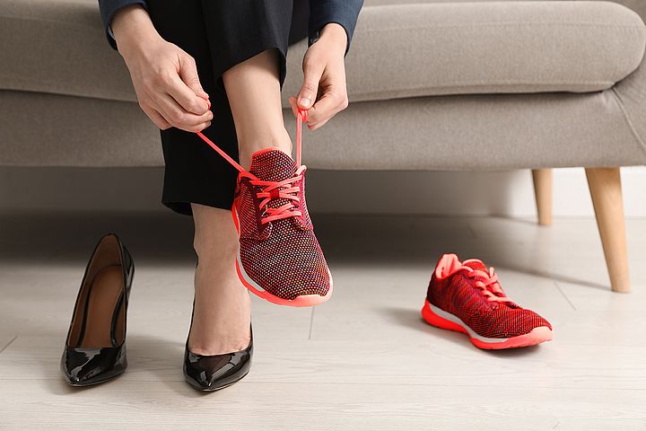 Eine Person sitzt auf einem hellgrauen Sofa und zieht einen roten Sportschuh an, während sie auf dem anderen Fuß noch schwarze High Heels trägt. Der zweite Sportschuh sowie der zweite High Heel liegen daneben auf dem Boden.