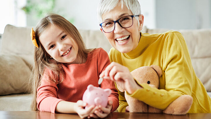 Sehr moderne Oma mit Brille und Kurzhaarschnitt wirft Geld in das Sparschwein ihrer Enkeltochter.