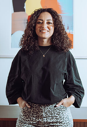 Foto von einer selbstbewussten jungen Frau mit lockigem dunklem Haar. Sie trägt einen schwarzes, langärmliges Oberteil und eine Hose im Animal Print, dahinter ist ein einrahmtes buntes Bild an der Wand zu sehen.