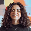 Foto von einer selbstbewussten jungen Frau mit lockigem dunklem Haar. Sie trägt einen schwarzes, langärmliges Oberteil und eine Hose im Animal Print, dahinter ist ein einrahmtes buntes Bild an der Wand zu sehen.