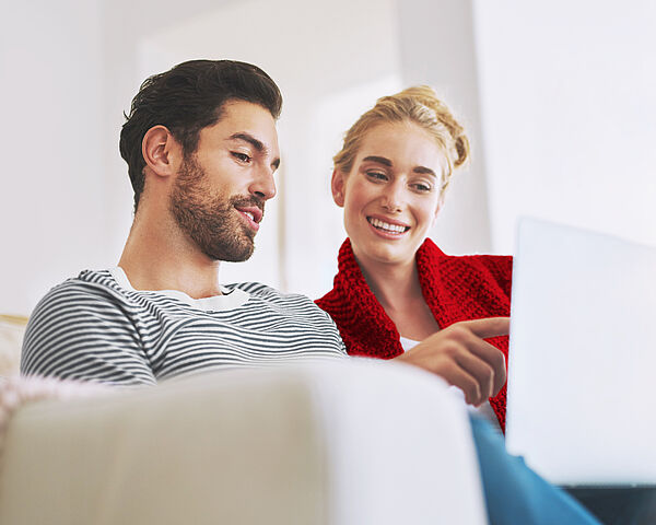 Ein junger Mann sitzt mit seiner Freundin auf der Couch und zeigt ihr etwas auf seinem Laptop.