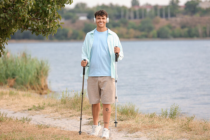 Ein junger Mann in kurzer Hose und hellblauem T-Shirt steht auf einem Weg am Wasser, hält Nordic-Walking-Stöcke in den Händen und hat weiße Kopfhörer auf.