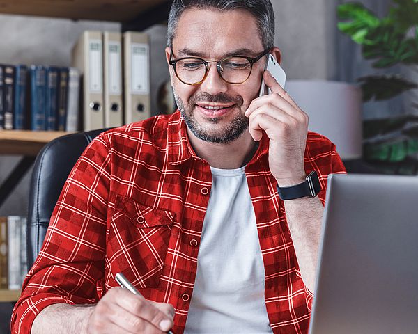 Ein Mann sitzt am Laptop im Büro und schreibt etwas auf einem Notizblock während er telefoniert.