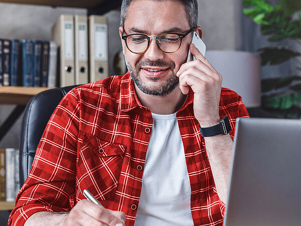 Ein Mann sitzt am Laptop im Büro und schreibt etwas auf einem Notizblock während er telefoniert.