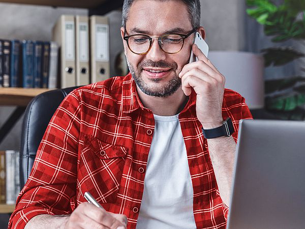 Ein Mann sitzt am Laptop im Büro und schreibt etwas auf einem Notizblock während er telefoniert.