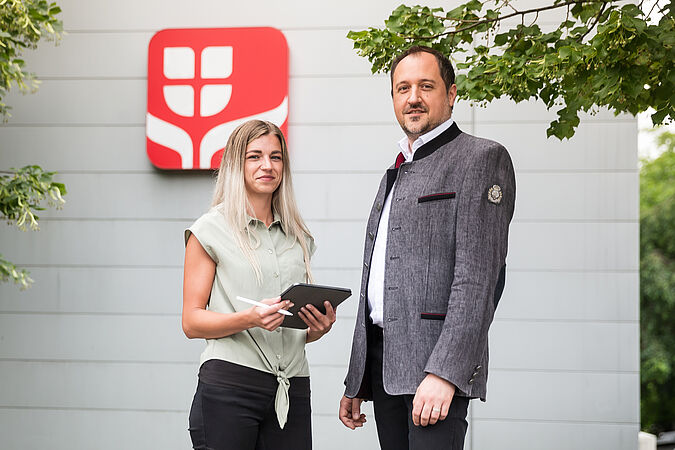 Eine blonde Frau und ein dunkelhaariger Mann stehen vor dem Logo der Wiener Städtischen und lächeln.