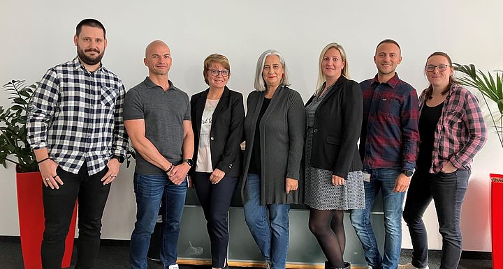 Gruppenfoto (7 Personen) vor einer weißen Wand in einem Büro.