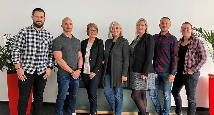 Gruppenfoto (7 Personen) vor einer weißen Wand in einem Büro.