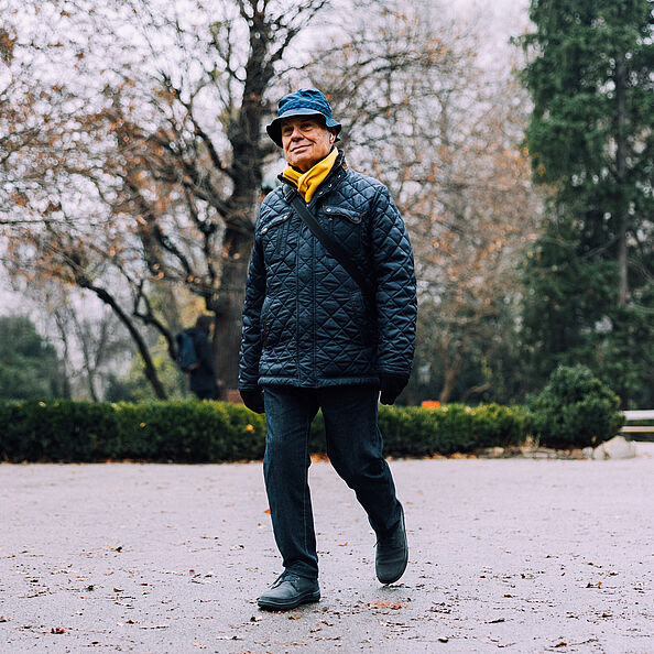 Ein älterer Mann in dunkler winterlicher Kleidung und Fischerhut spaziert im Park, im Hintergrund sind Bäume zu sehen.