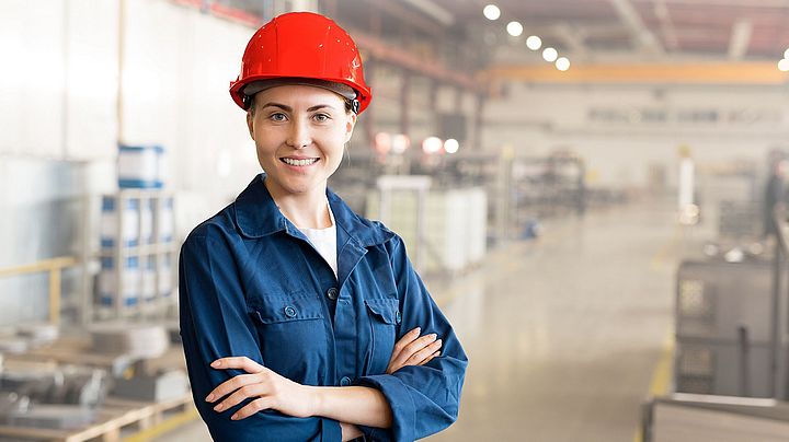 Eine lächelnde junge Frau trägt einen Helm und steht in einer Fabrik.