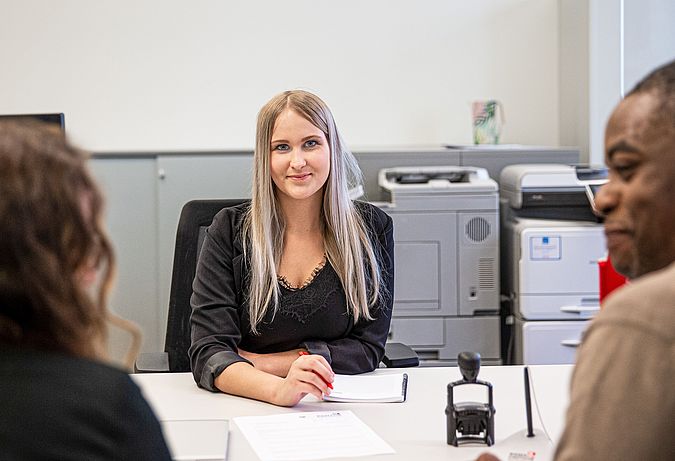 Blonde Dame sitzt an einem Schreibtisch. Ihr gegenüber sitzen ein Mann und eine Frau. Sie unterhalten sich.