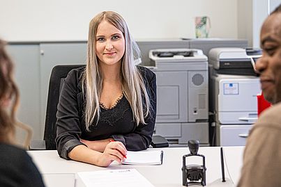 Blonde Dame sitzt an einem Schreibtisch. Ihr gegenüber sitzen ein Mann und eine Frau. Sie unterhalten sich.