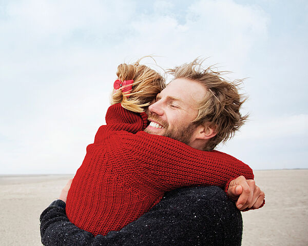 Eine junge Frau mit hochgesteckten, blonden Haaren und roten Pullover umarmt einen jungen, blonden Mann mit schwarzem Pullover. Es ist windig und sie stehen an einem Strand.