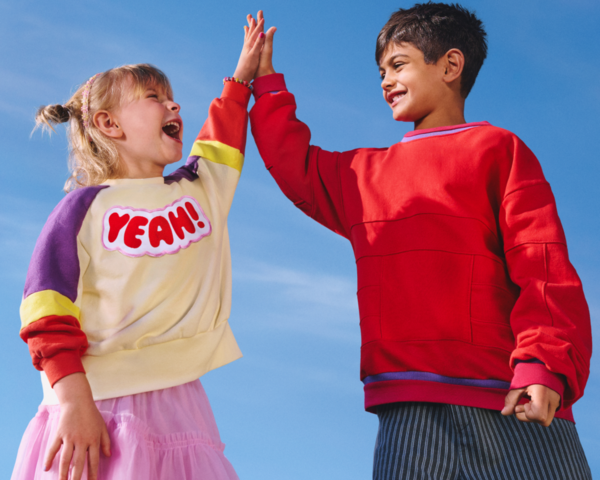 Zwei lachende Kinder geben sich vor blauem Himmel ein High Five. Ein Kind trägt einen bunten Pullover mit der Aufschrift ‚YEAH!‘, das andere einen roten Pullover.