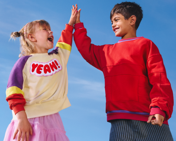 Zwei lachende Kinder geben sich vor blauem Himmel ein High Five. Ein Kind trägt einen bunten Pullover mit der Aufschrift ‚YEAH!‘, das andere einen roten Pullover.
