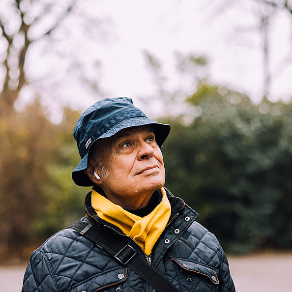 Porträtfoto von einem älteren Mann im Park - er trägt einen Fischerhut, eine dunkle Jacke, einen gelben Schal und blickt zur Seite..
