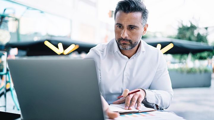 Ein Geschäftsmann sitzt am Schreibtisch vor seinem Laptop und macht sich konzentriert Notizen