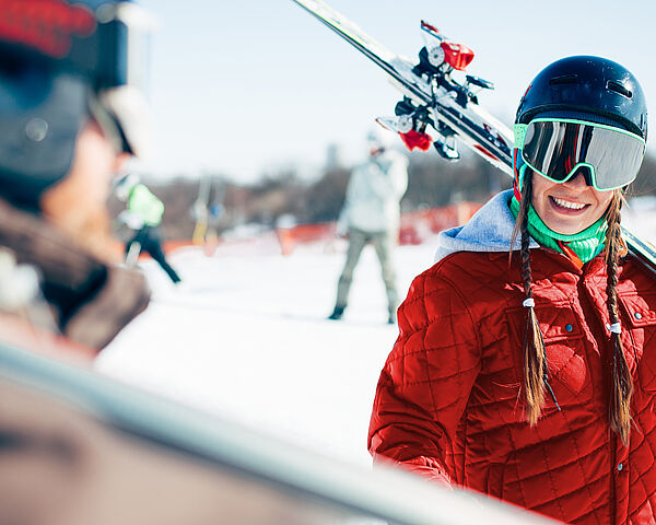 Eine Frau befindet sich mit einem Mann in Skibekleidung auf einer Skipiste, wobei beide Skier auf den Schultern tragen. Die Frau lächelt dabei den Mann an.  