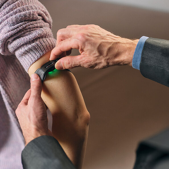 Zwei Männerhände befestigen eine Smartwatch am Oberarm einer Frau mit blasslila Strickjacke.