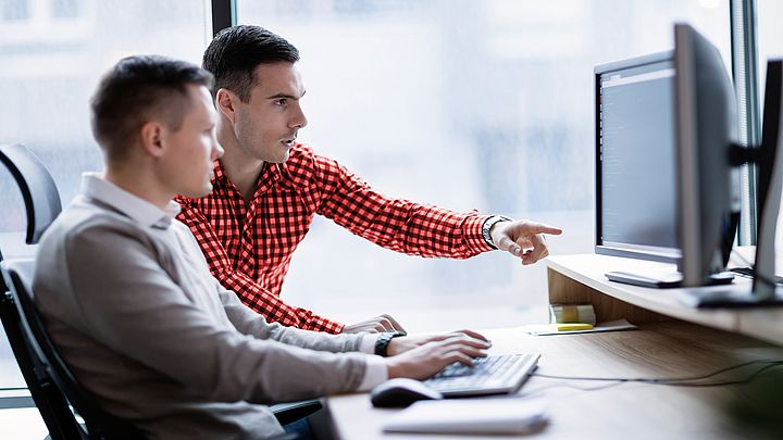 Zwei Geschäftsmänner sitzen in einem Büro und arbeiten gemeinsam fokussiert am Computer.