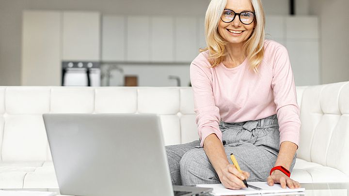 Eine Frau sitzt lächelnd auf einem Sofa vor ihrem Laptop und schreibt Notizen auf ihrem Notizblock.