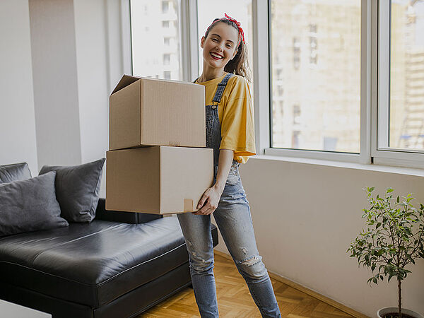 Mädchen steht lächelnd mit zwei Umzugskartons in der Hand in der ersten eigenen Wohnung.