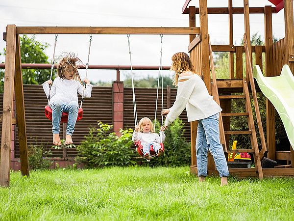 Zwei Kinder befinden sich auf einem Spielplatz, wo sie beide auf einer Schaukel sitzen und von ihrer Mutter angestoßen werden