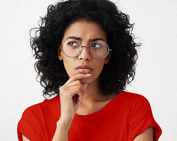 Eine Frau, mit lockigem Haar und Brille, fasst sich an das Kinn und setzt einen nachdenklichen Blick auf 