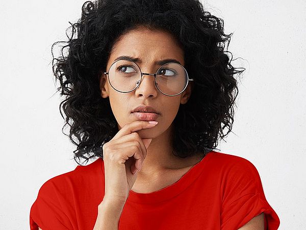 Eine Frau, mit lockigem Haar und Brille, fasst sich an das Kinn und setzt einen nachdenklichen Blick auf 