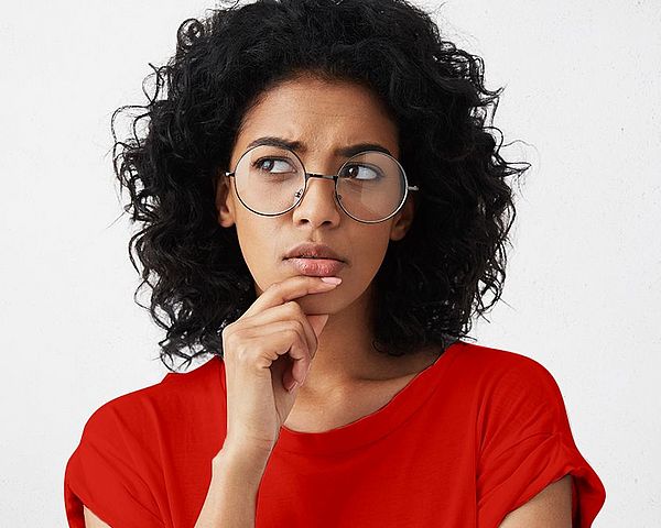 Eine Frau, mit lockigem Haar und Brille, fasst sich an das Kinn und setzt einen nachdenklichen Blick auf 