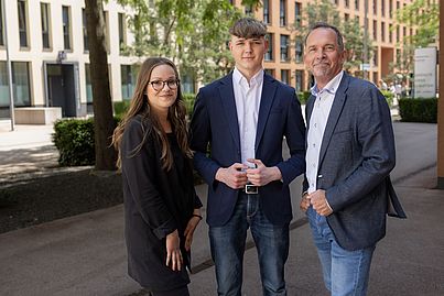 Eine junge Dame mit Brille und zwei Herren im Anzug stehen auf einem Gehsteig nebeneinander.