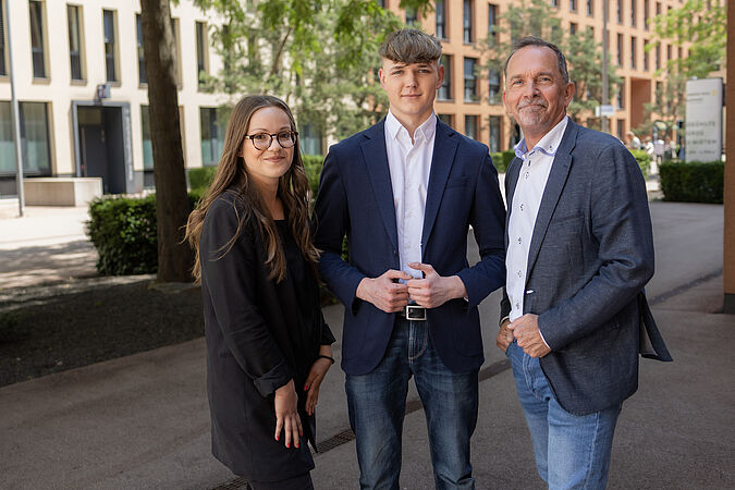 Eine junge Dame mit Brille und zwei Herren im Anzug stehen auf einem Gehsteig nebeneinander.