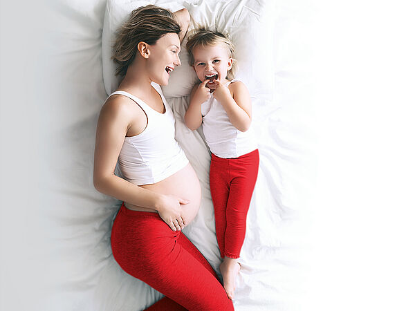 Eine schwangere Mutter liegt mit ihrem Kind lachend im Bett.