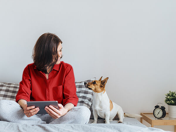 Eine junge Frau sitzt lächelnd mit ihrem Tablet und ihrem Hund im Bett.