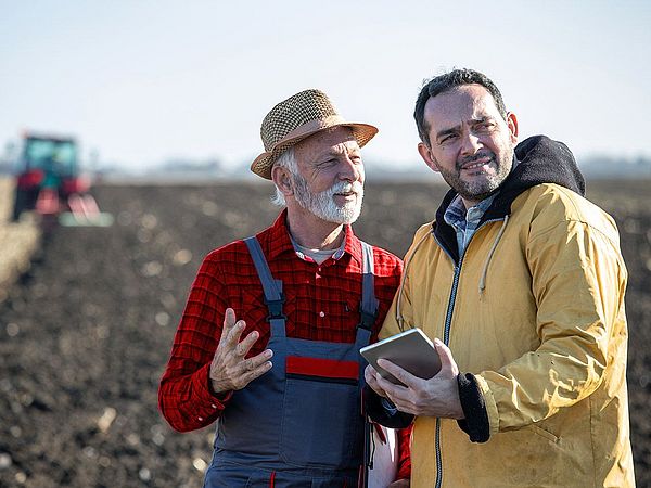 Ein Landwirt und ein Mann mit Tablet besprechen sich auf einem Feld. Dabei befindet sich im Hintergrund ein Traktor. 