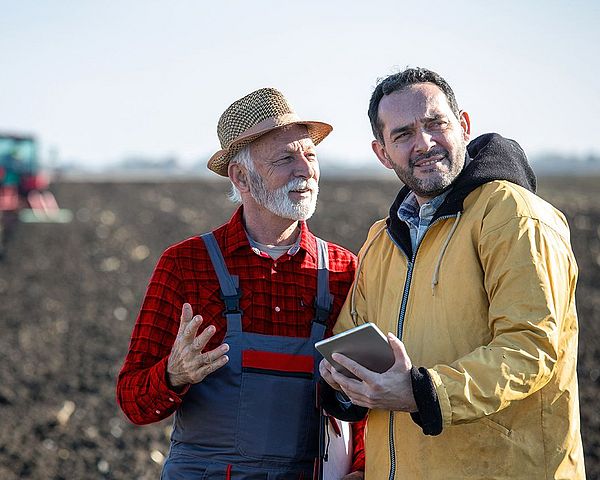 Ein Landwirt und ein Mann mit Tablet besprechen sich auf einem Feld. Dabei befindet sich im Hintergrund ein Traktor. 